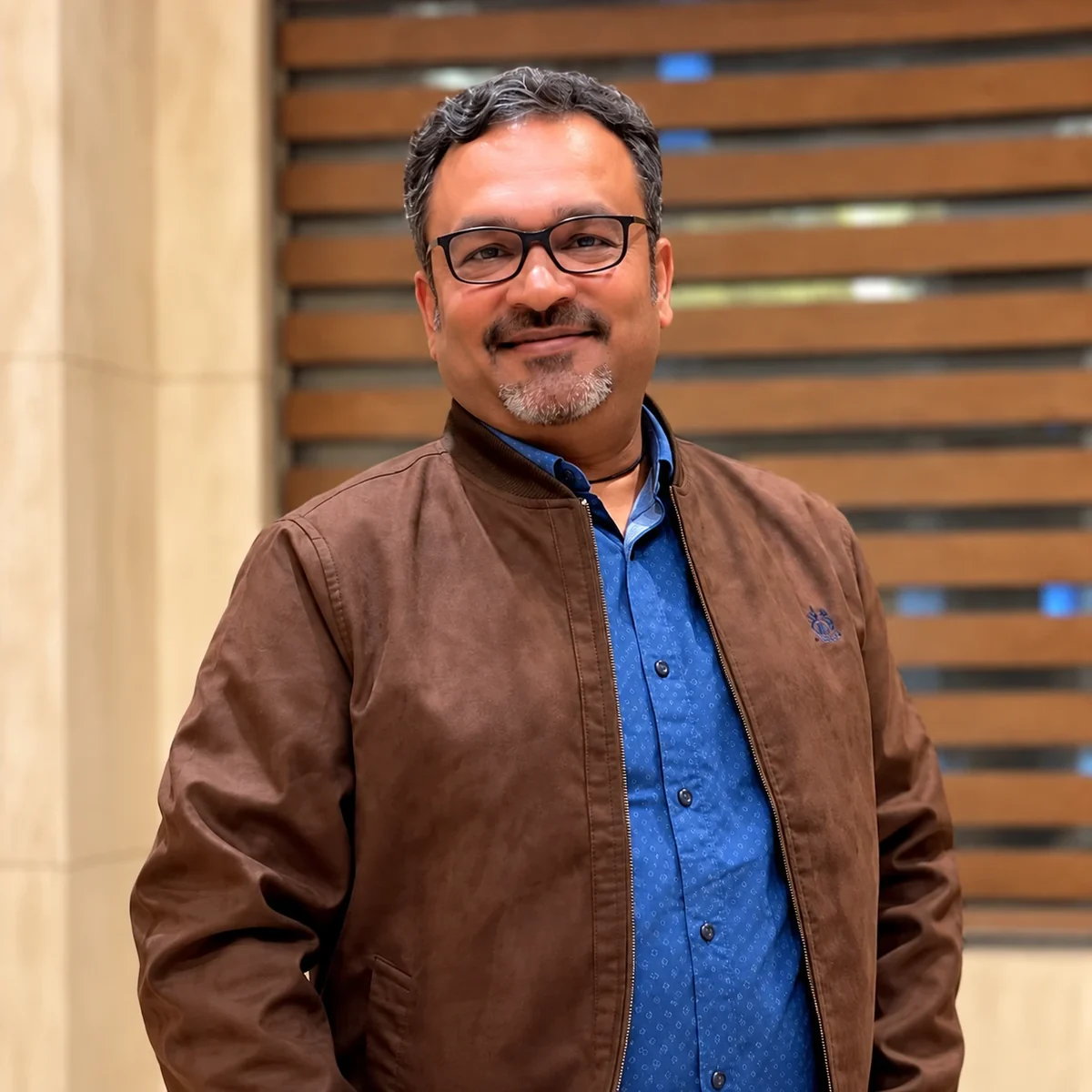 Amit Govil, founder and CEO of XONIK, smiling, in glasses and a brown jacket over a blue shirt.